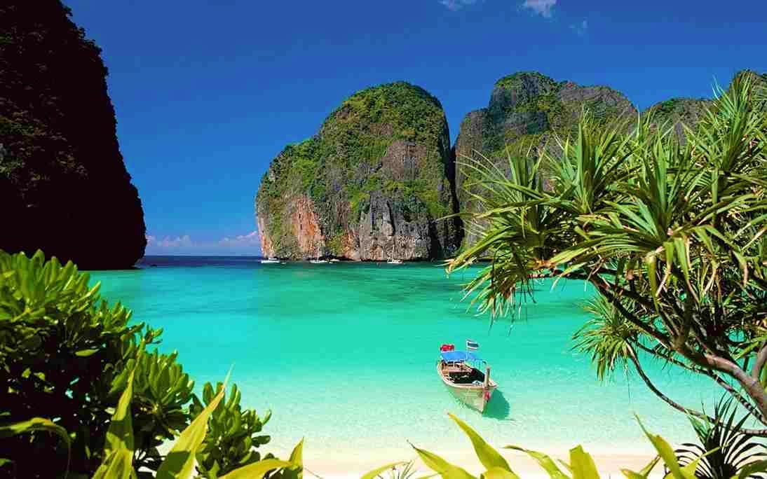 Tropical beach cove with turquoise water, limestone cliffs, longtail boat, and palm trees framing the pristine sandy shore